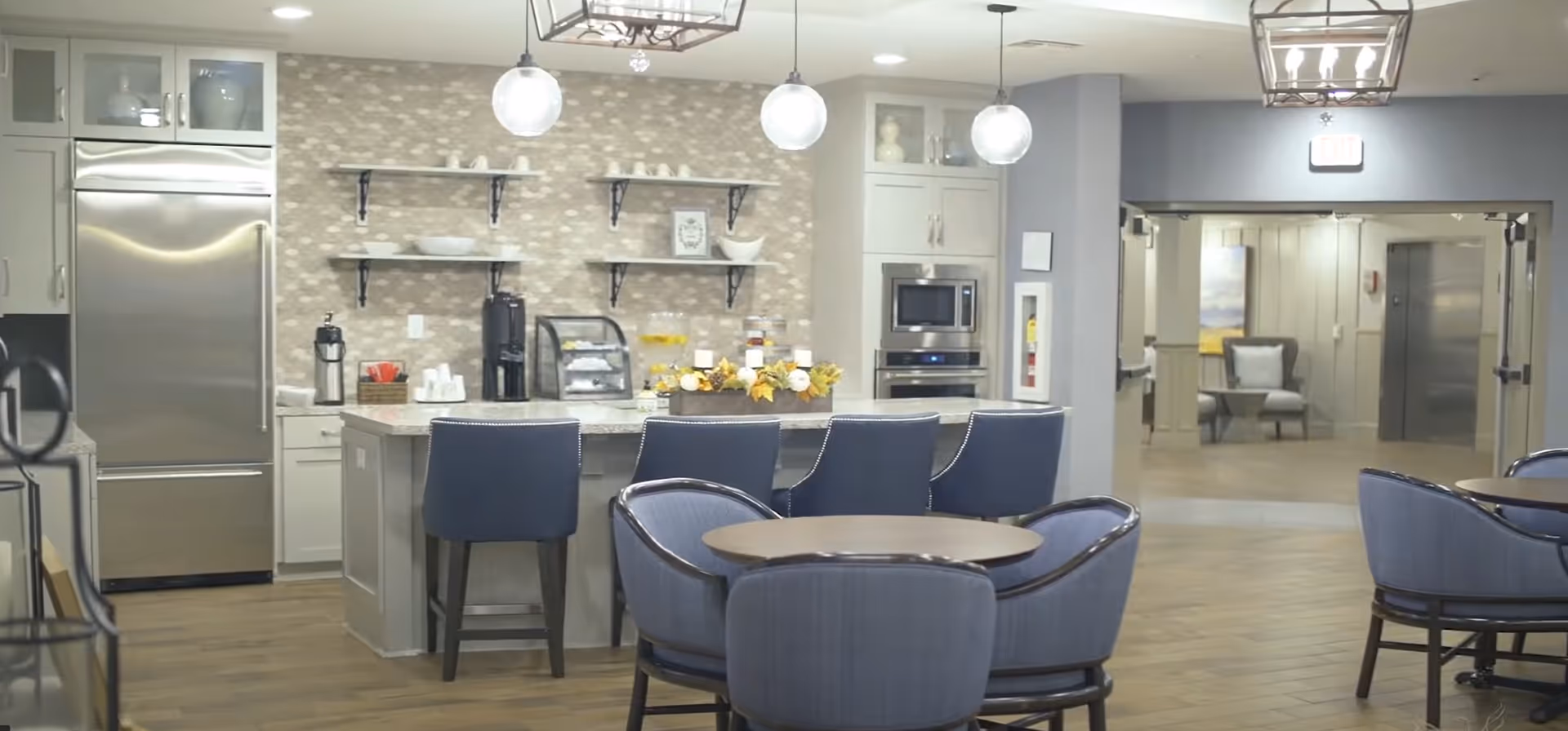 A modern kitchen and dining area in a senior living facility with a large stainless steel refrigerator, a kitchen island with four blue bar stools, pendant lights hanging from the ceiling, open shelves with decorative items, and round tables with blue upholstered chairs. The background shows a hallway with seating and an elevator.