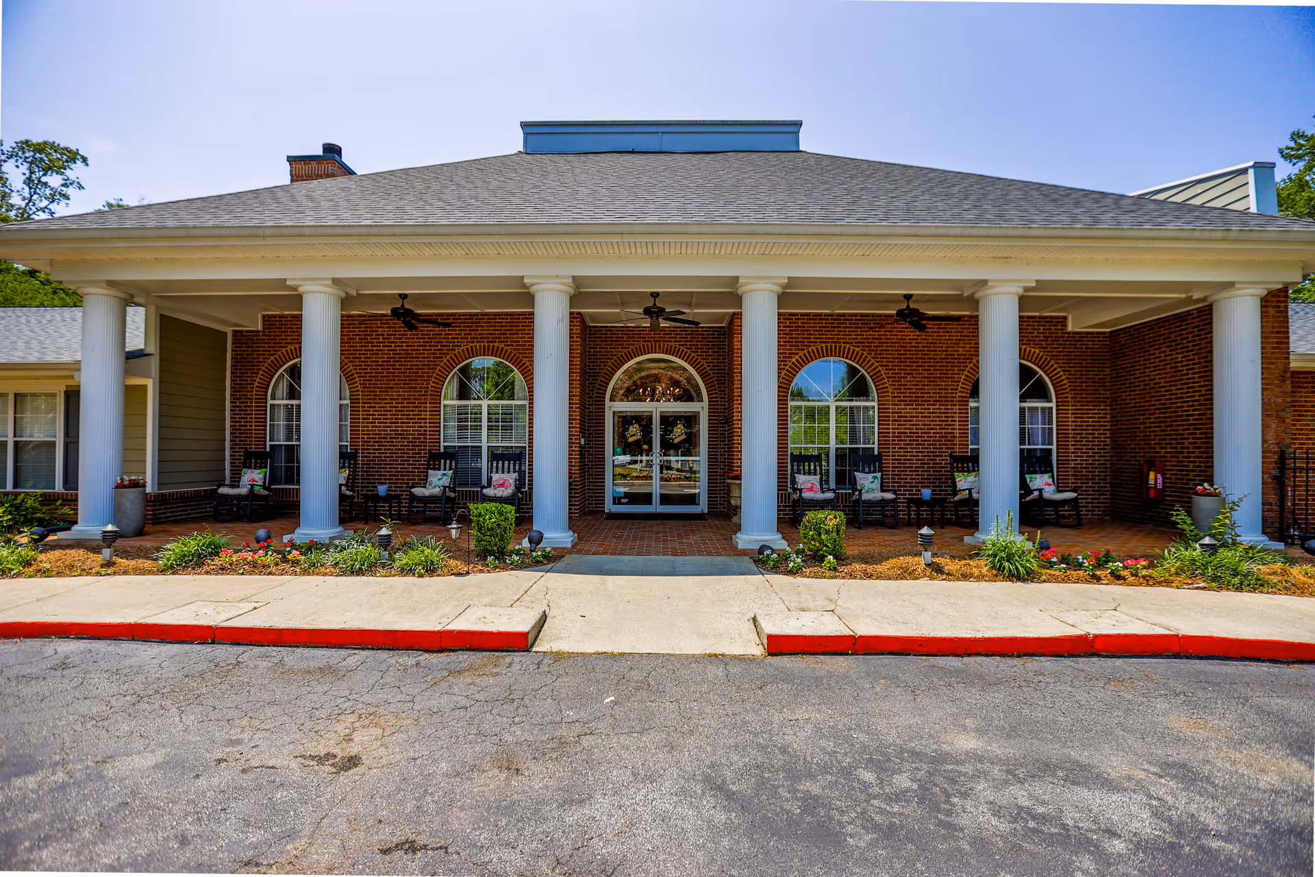 Front exterior view of a brick building with white columns supporting a covered entrance. There are several rocking chairs with cushions on the porch, flower beds with plants and flowers along the walkway, and a clear blue sky above.