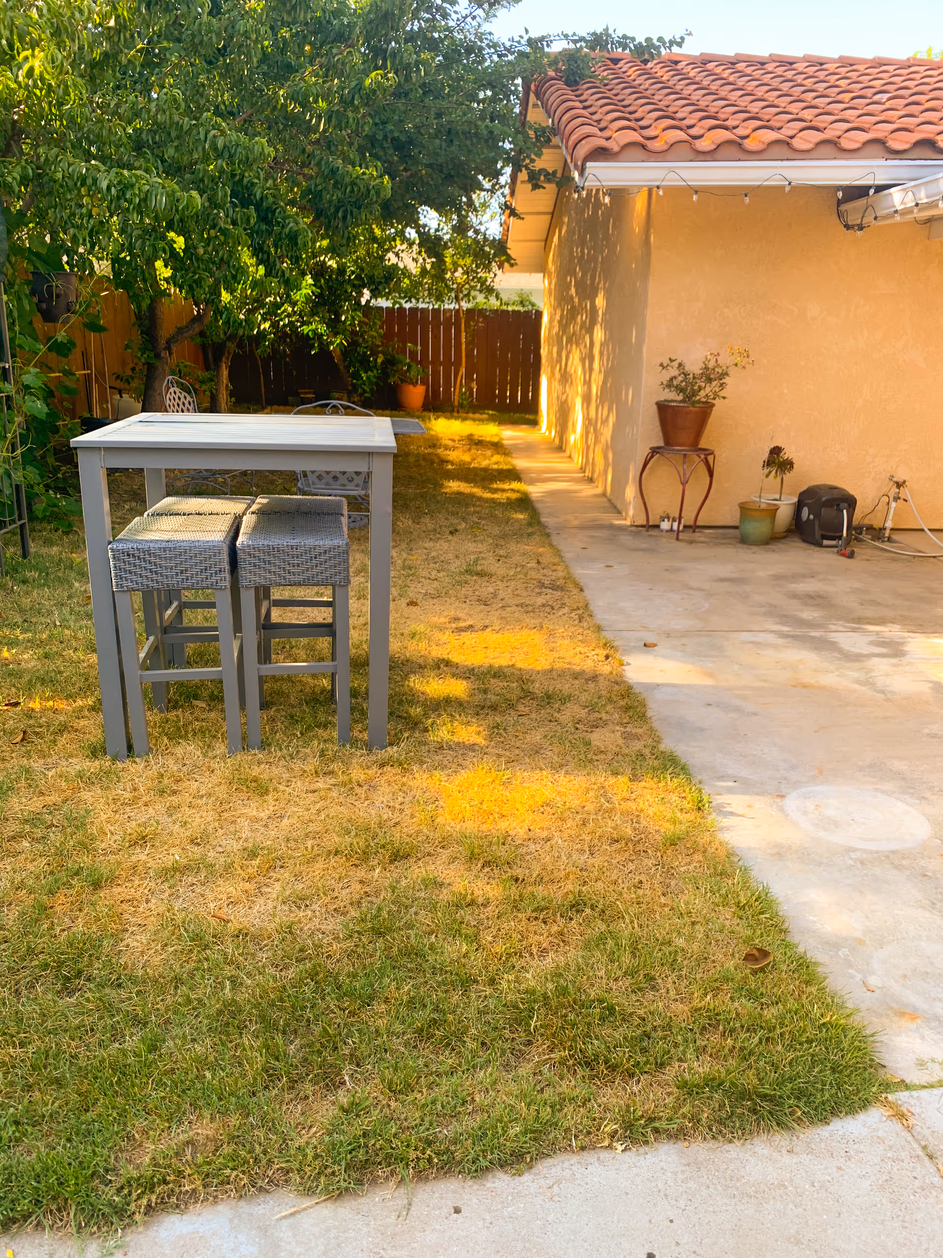Outdoor backyard area with a small gray table and two matching stools on a patchy grass lawn. There is a concrete patio area adjacent to a beige stucco building with a red tile roof. Several potted plants are placed on the patio and a wooden fence and green trees are visible in the background.