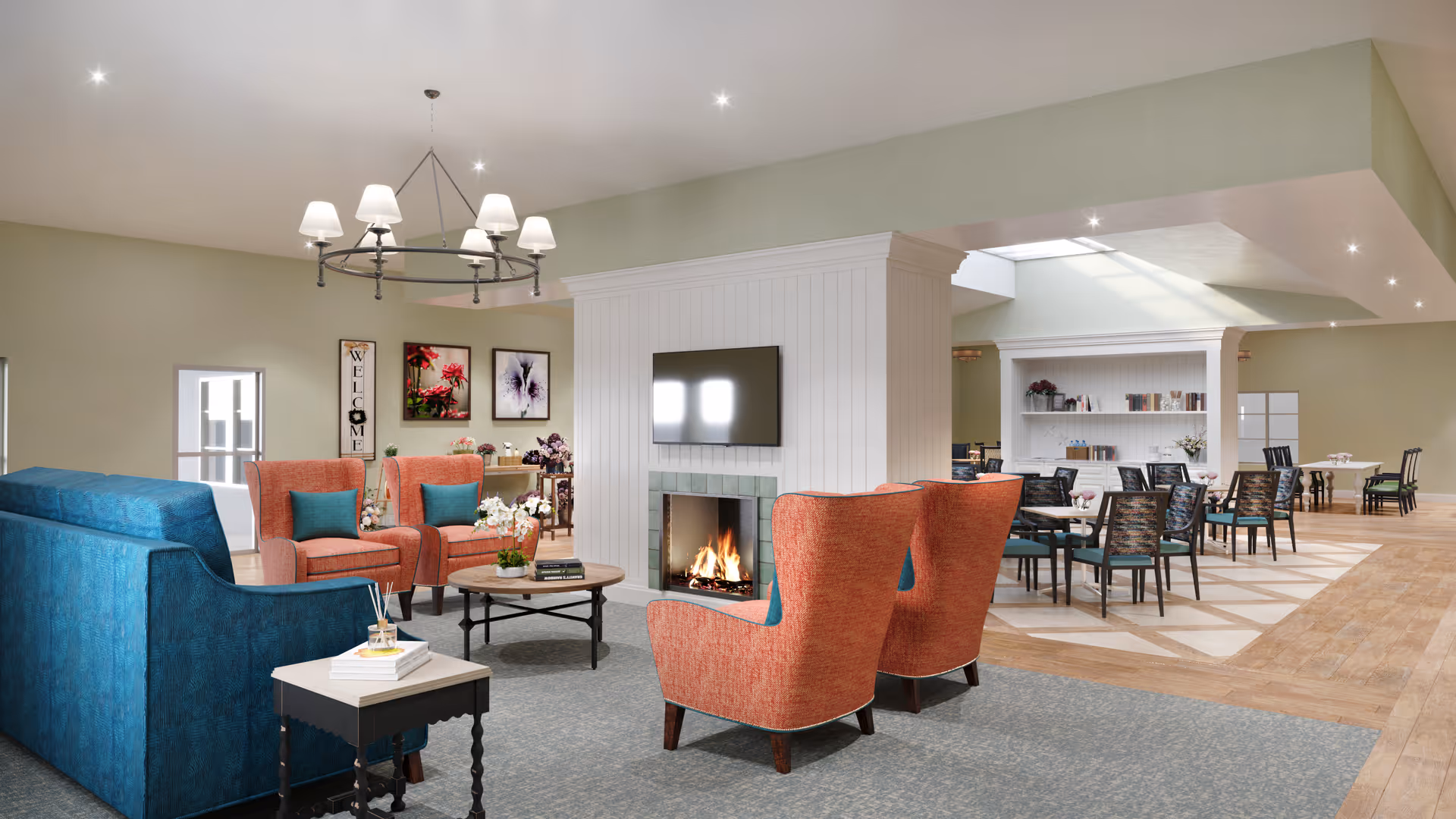 A spacious senior living common area featuring a cozy seating arrangement with blue and orange armchairs around a coffee table in front of a fireplace with a mounted TV above it. In the background, there is a dining area with tables and chairs, bookshelves, and soft lighting from ceiling fixtures.