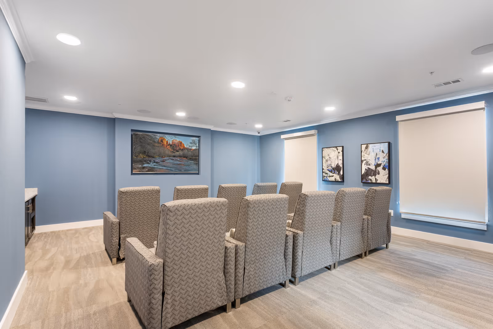 A small theater room with two rows of gray patterned recliner chairs facing a large flat-screen TV mounted on a blue wall. The room has beige carpet, recessed ceiling lights, and two windows with white roller shades. There are two framed artworks on the right wall.