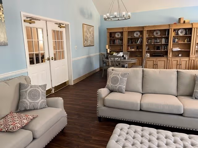 A bright common area with light-gray sofas, a tufted ottoman, wooden bookcases, a dining table, and double French doors.