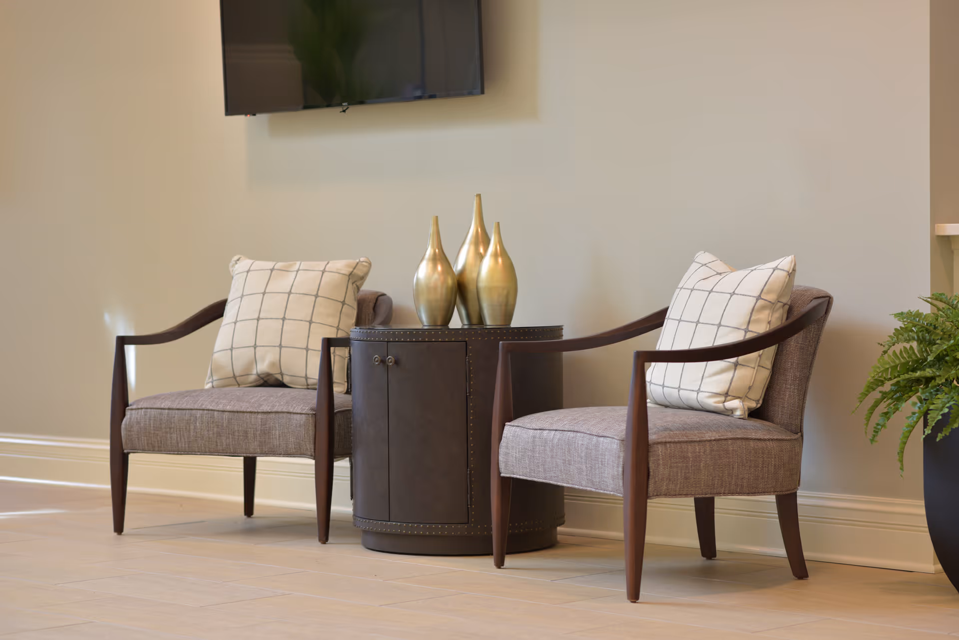 Two upholstered armchairs with checked pillows flank a round side table topped with three metallic vases in a tidy interior seating area.