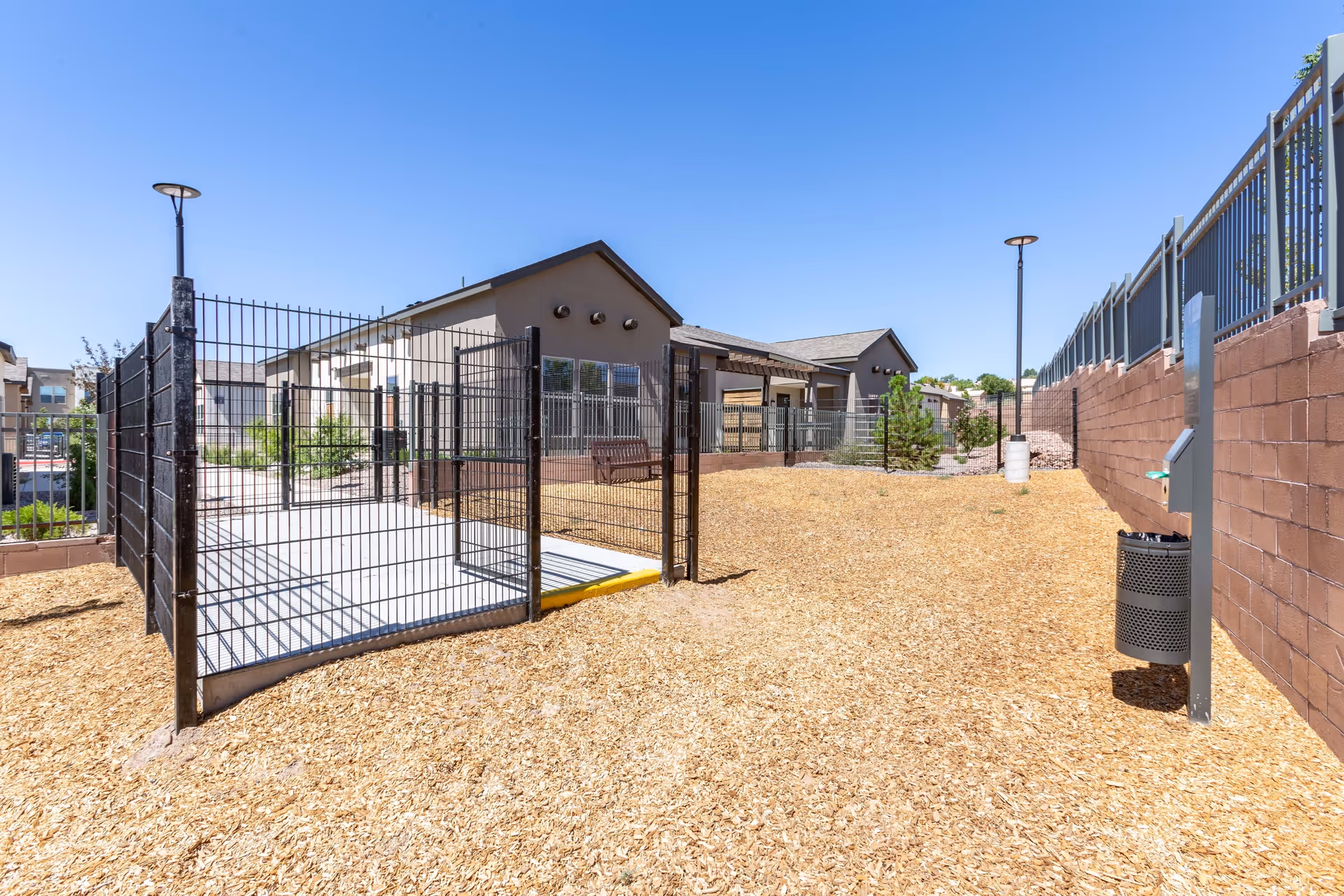 A fenced outdoor courtyard covered in wood chips with a gated walkway, lamp posts, a trash receptacle, and low single-story buildings in the background.