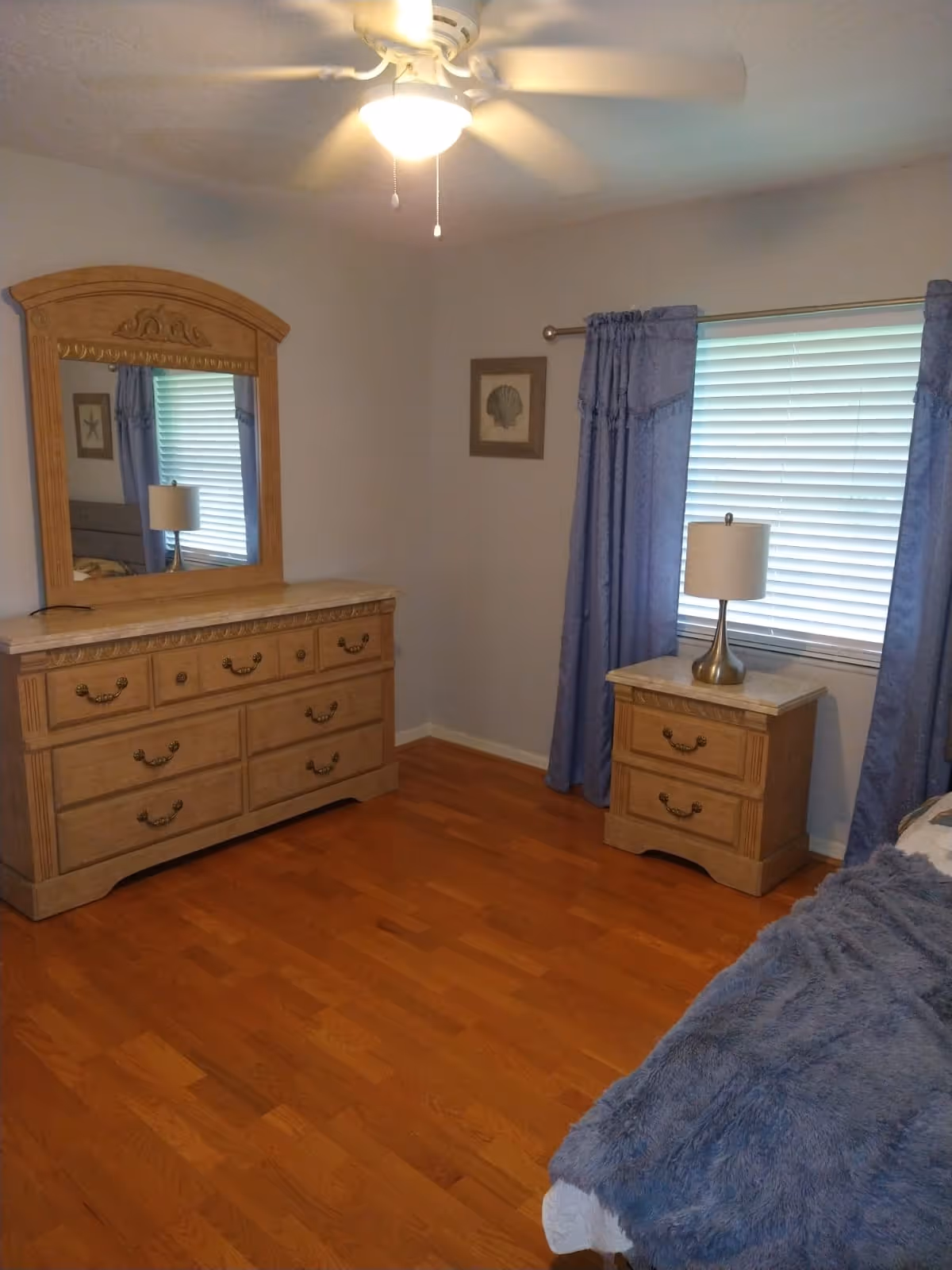 A bedroom with wooden flooring, a wooden dresser with a large mirror, a nightstand with a lamp, a window with blue curtains, and part of a bed with a blue blanket. The ceiling has a white fan with a light.