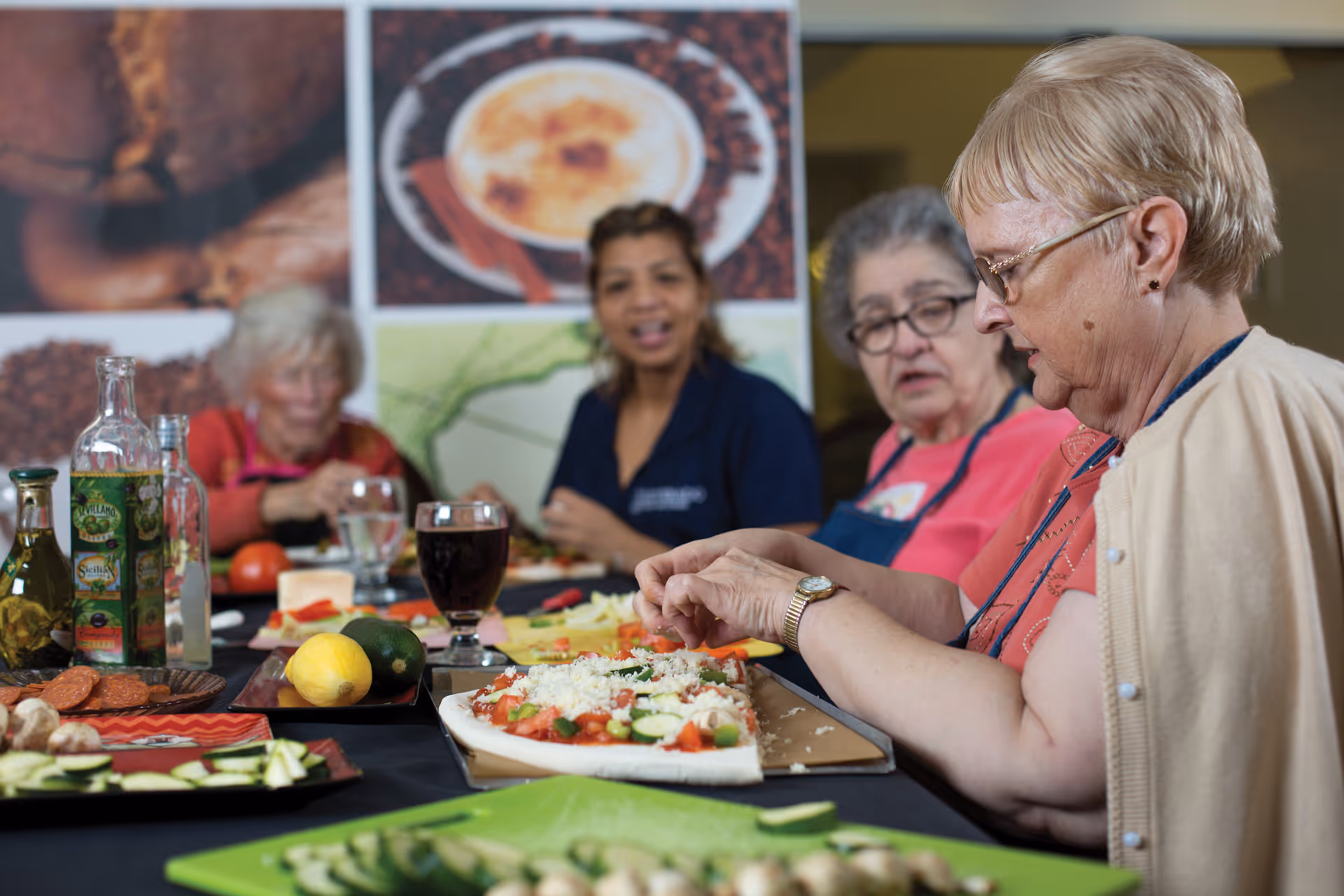 A group of elderly women and a caregiver sitting around a table preparing food together. The table is filled with various ingredients including sliced vegetables, bottles of oil, and a partially assembled pizza. The background features large images of coffee and spices.