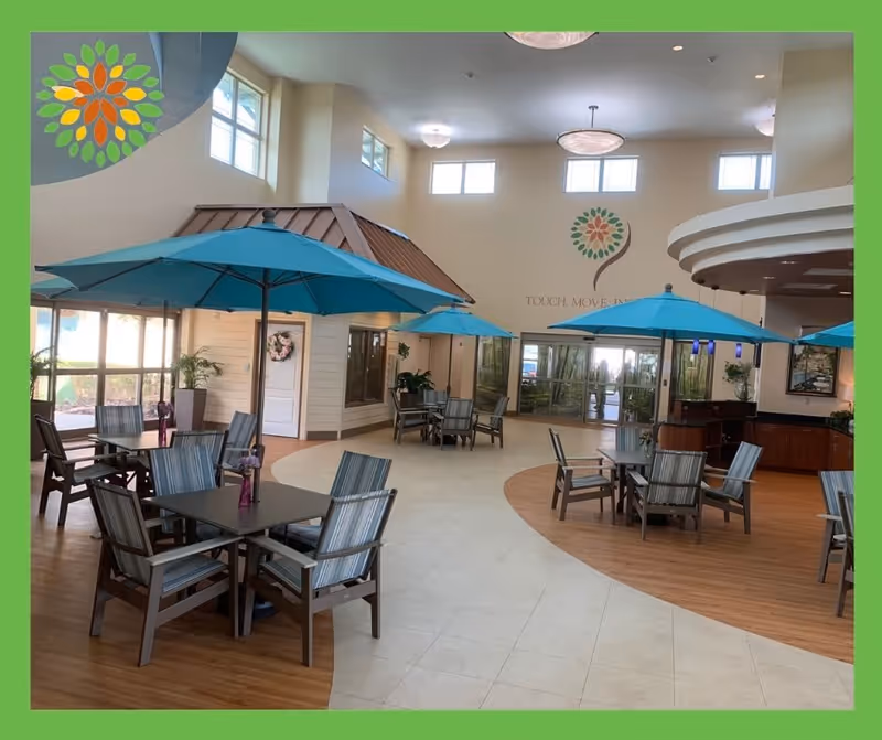 Spacious indoor common area with multiple tables and chairs arranged under blue umbrellas. The room has high ceilings with several windows allowing natural light, and a decorative logo on the far wall with the words 'TOUCH. MOVE. RAISE.'. The floor is a combination of wood and tile, and there are plants and a small structure resembling a house inside the room.