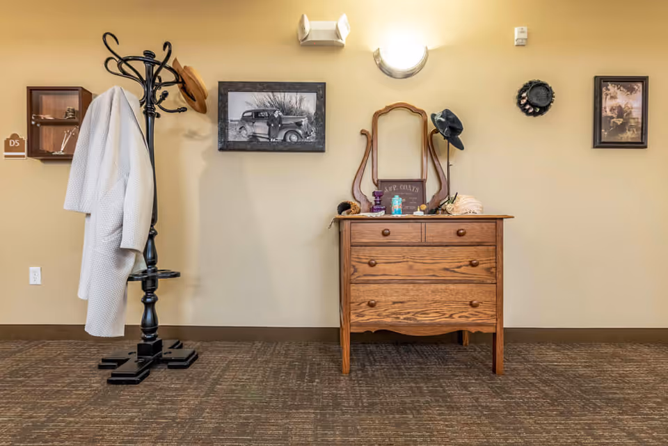 Interior view of a room with a wooden dresser topped with a mirror, hats, and decorative items. To the left is a black coat rack holding a white coat and a brown hat. The beige wall behind features framed black and white photographs and a wall light fixture above the dresser.