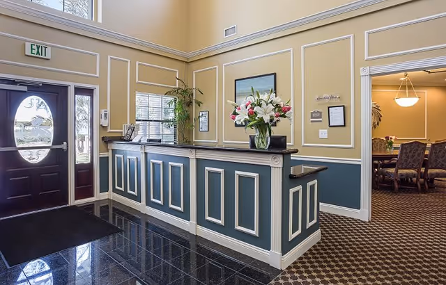 Reception lobby with a paneled front desk, vase of lilies, decorative wall trim, and an adjacent dining area visible through a doorway.