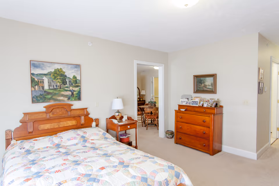 Sunlit bedroom with a wooden headboard bed covered by a patchwork quilt, a nightstand with a lamp, a wooden dresser with framed photos, and an open doorway into a dining area.