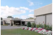 Exterior view of a single-story building with a flat roof, surrounded by a well-maintained lawn and flower beds with white and pink flowers under a partly cloudy sky.