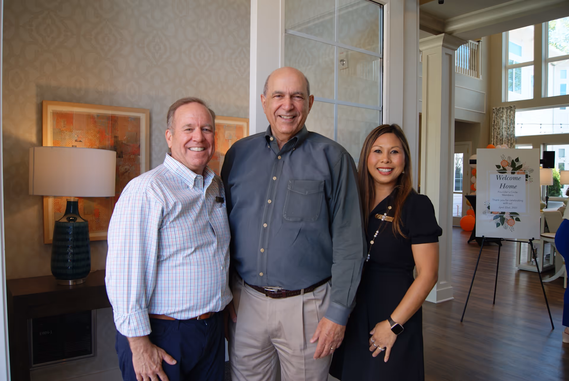 Three smiling adults stand together in a bright facility lobby beside a lamp and an easel-mounted "Welcome Home" sign.