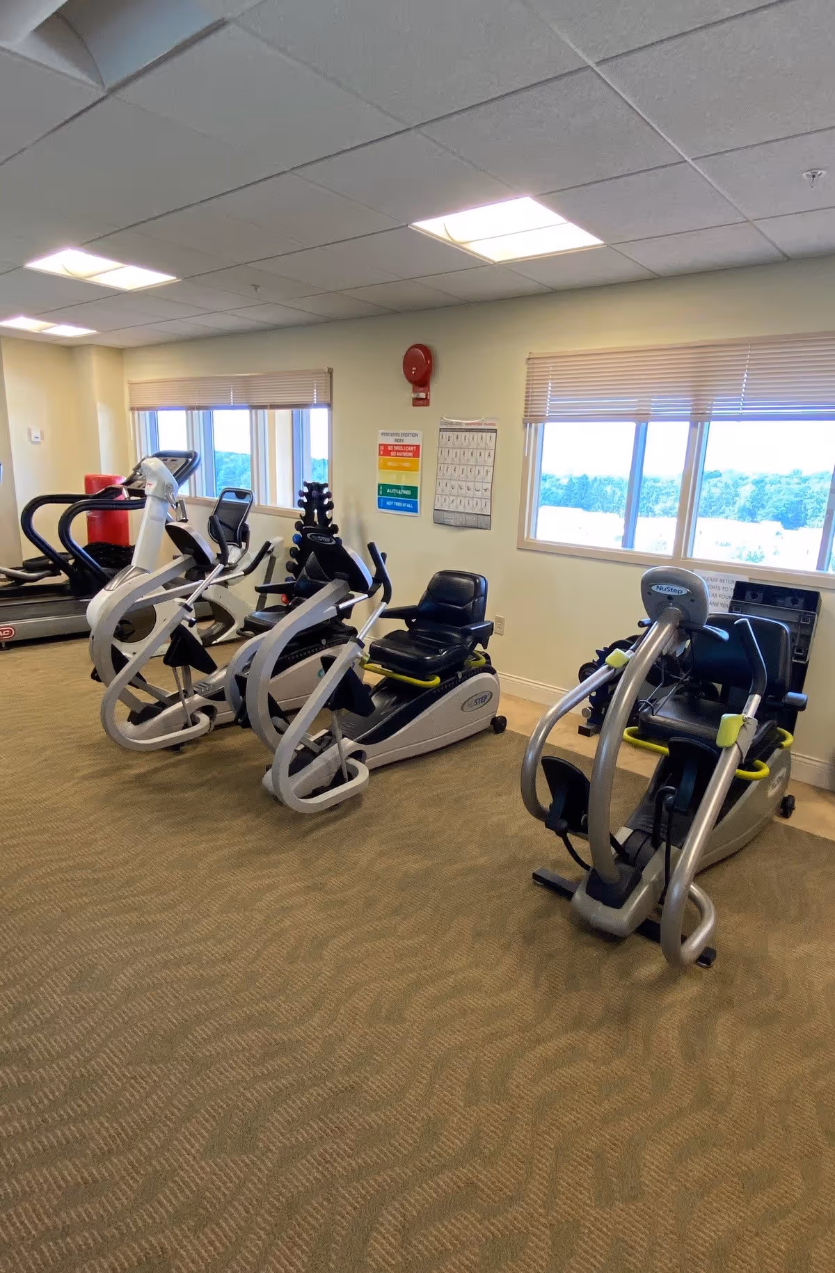 A fitness room with several exercise machines including recumbent bikes and a treadmill, beige carpeted floor, large windows with blinds, and a wall-mounted fire alarm and informational posters.