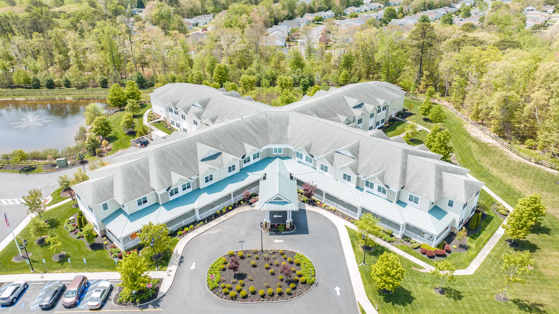 Aerial view of The Terraces at Seacrest Village senior living facility showing a large, two-story building with a circular driveway and landscaped garden in front. Surrounding the building are green lawns, trees, a pond with a fountain, and a residential neighborhood in the background.