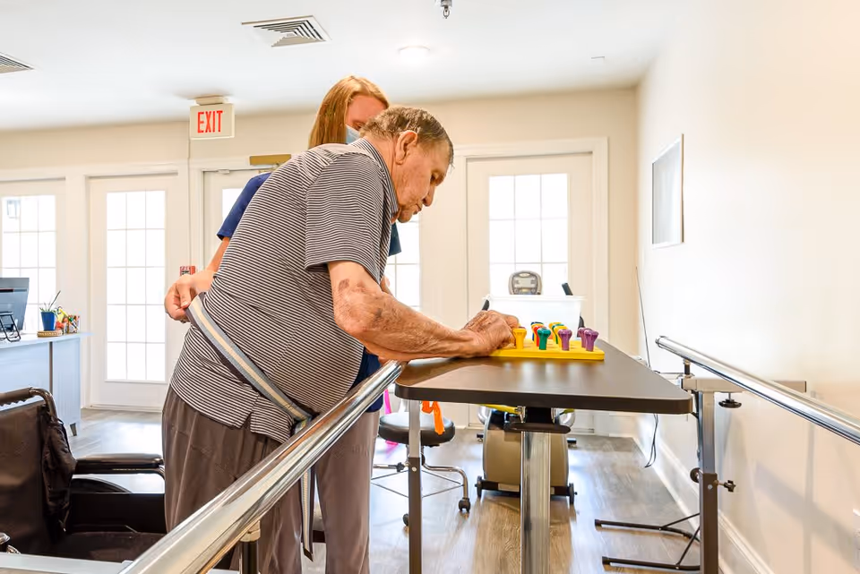 An elderly man wearing a striped shirt is engaged in a hand exercise activity using a pegboard on a table, assisted by a caregiver in a bright room with handrails and a wheelchair nearby.
