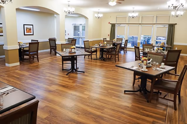 A spacious dining room with wooden floors, multiple tables set with cups, plates, and condiments, surrounded by chairs. The room has cream-colored walls, chandeliers, ceiling fans, and large windows with curtains letting in natural light.