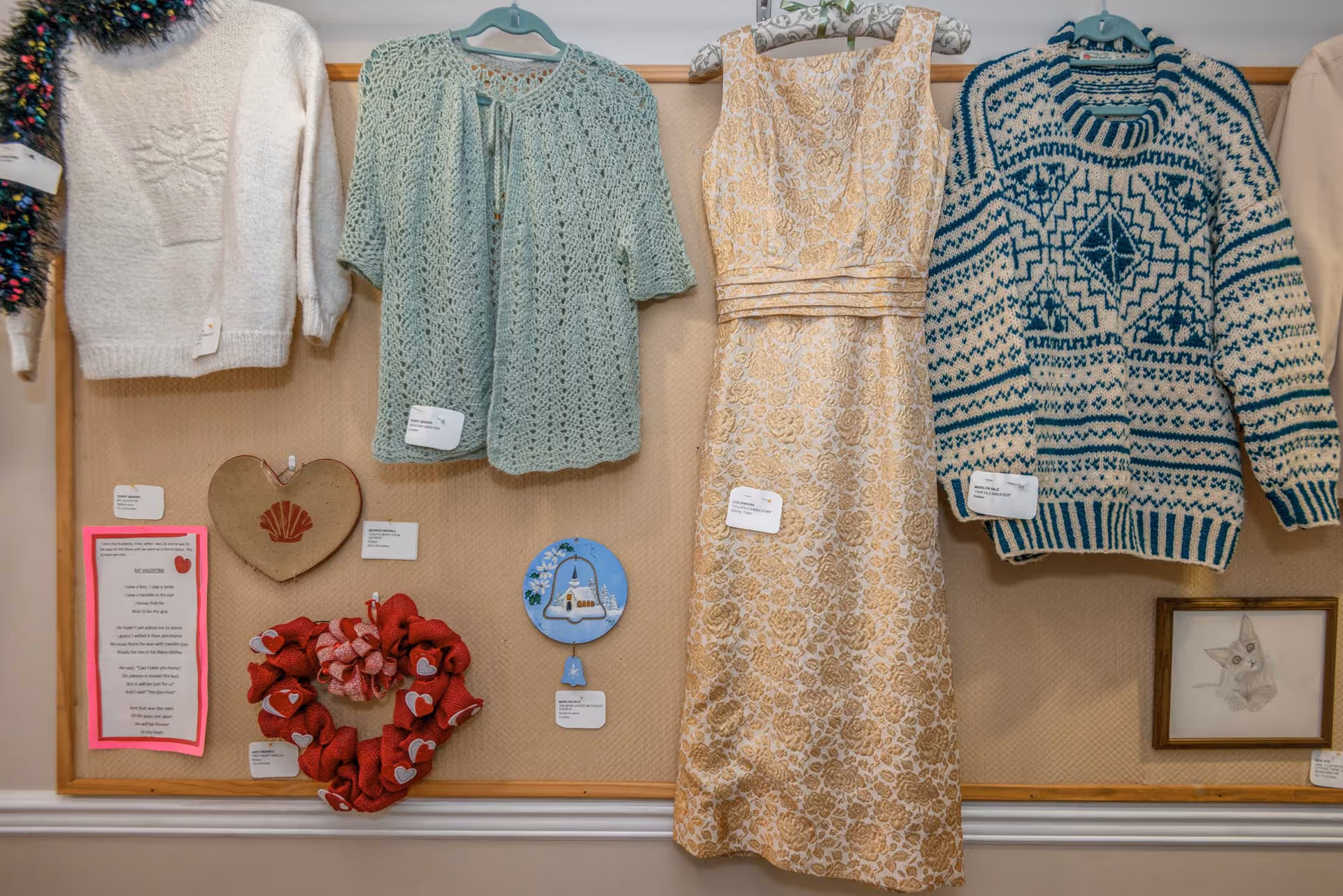 A display board featuring various handmade items including a white knitted sweater, a light green crocheted cardigan, a beige floral dress, and a blue and white patterned sweater. Below the clothing are decorative items such as a heart-shaped piece, a red wreath with hearts, a painted plate with a church, and a framed drawing of a cat. Each item has a small label attached.