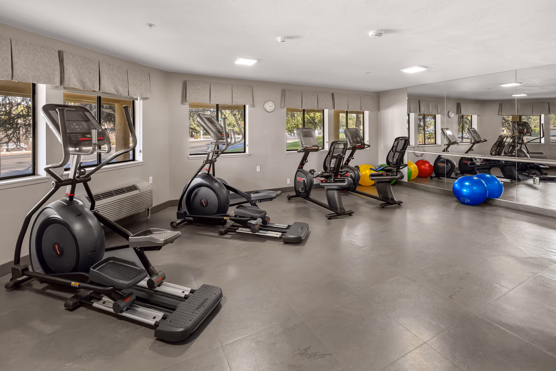 A fitness room with exercise equipment including two elliptical machines and two recumbent bikes. There are large windows with beige valances letting in natural light, a wall clock, and a large mirror covering one wall. Colorful exercise balls are placed near the mirror on the floor.