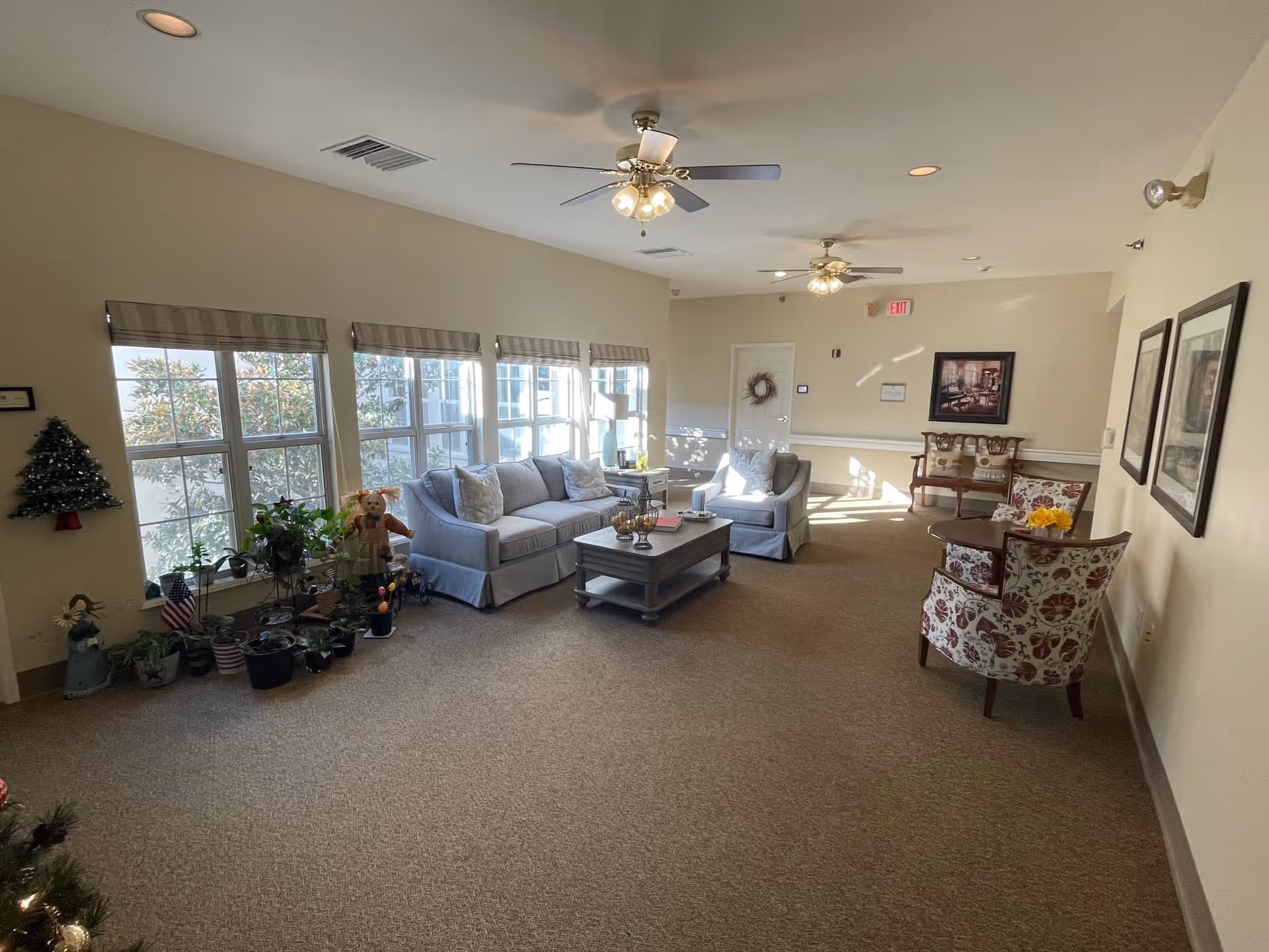 A bright and spacious living room area with beige walls and carpeted floor. The room features a light gray sofa and matching armchair around a wooden coffee table. There are multiple windows with blinds letting in natural light. Several potted plants and decorative items are placed near the windows. Two ceiling fans with lights hang from the ceiling. On the right side, there are two floral-patterned armchairs with a small round table between them, and framed pictures on the wall. A wooden bench and a door with a wreath are visible in the background.