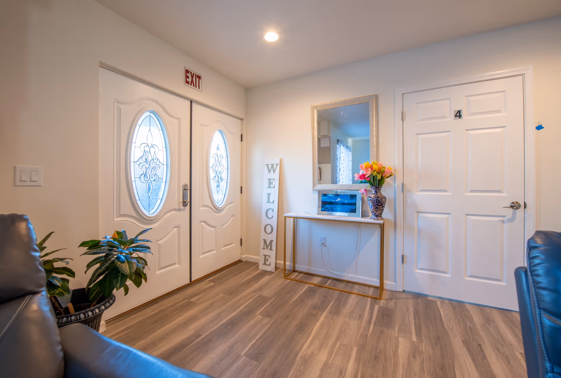 Bright foyer with double decorative glass doors, a 'WELCOME' sign, console table with mirror and flowers, and a numbered interior door.