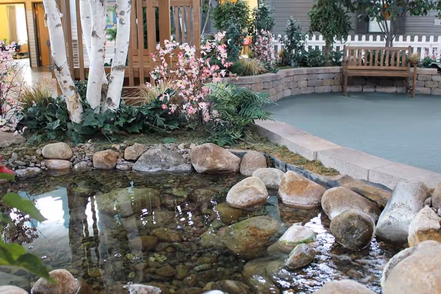 Indoor garden area featuring a small pond with clear water and rocks, surrounded by plants including white birch trees and pink flowering shrubs. There is a wooden bench and a white picket fence in the background.