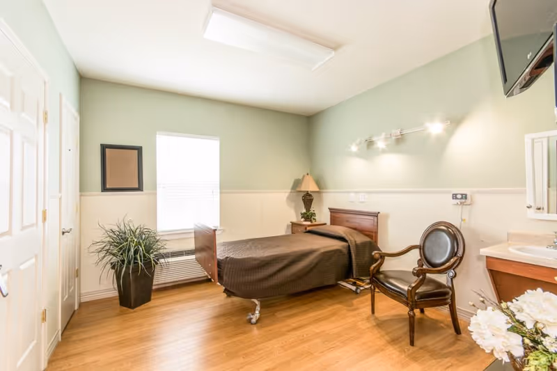 A clean and well-lit bedroom in a senior living facility with a single bed covered in a brown blanket, a wooden nightstand with a lamp and plant, a wooden chair with leather upholstery, a window with blinds, a potted plant on the floor, and a wall-mounted television. The room has light green walls with white wainscoting and wooden flooring.