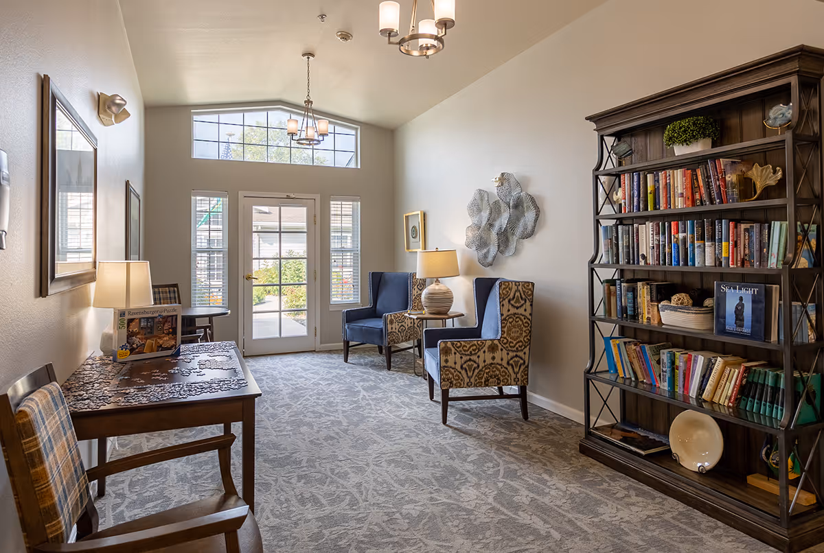 A bright seating area with bookshelves, patterned armchairs, a table with a lamp and puzzle, and glass doors leading outside.
