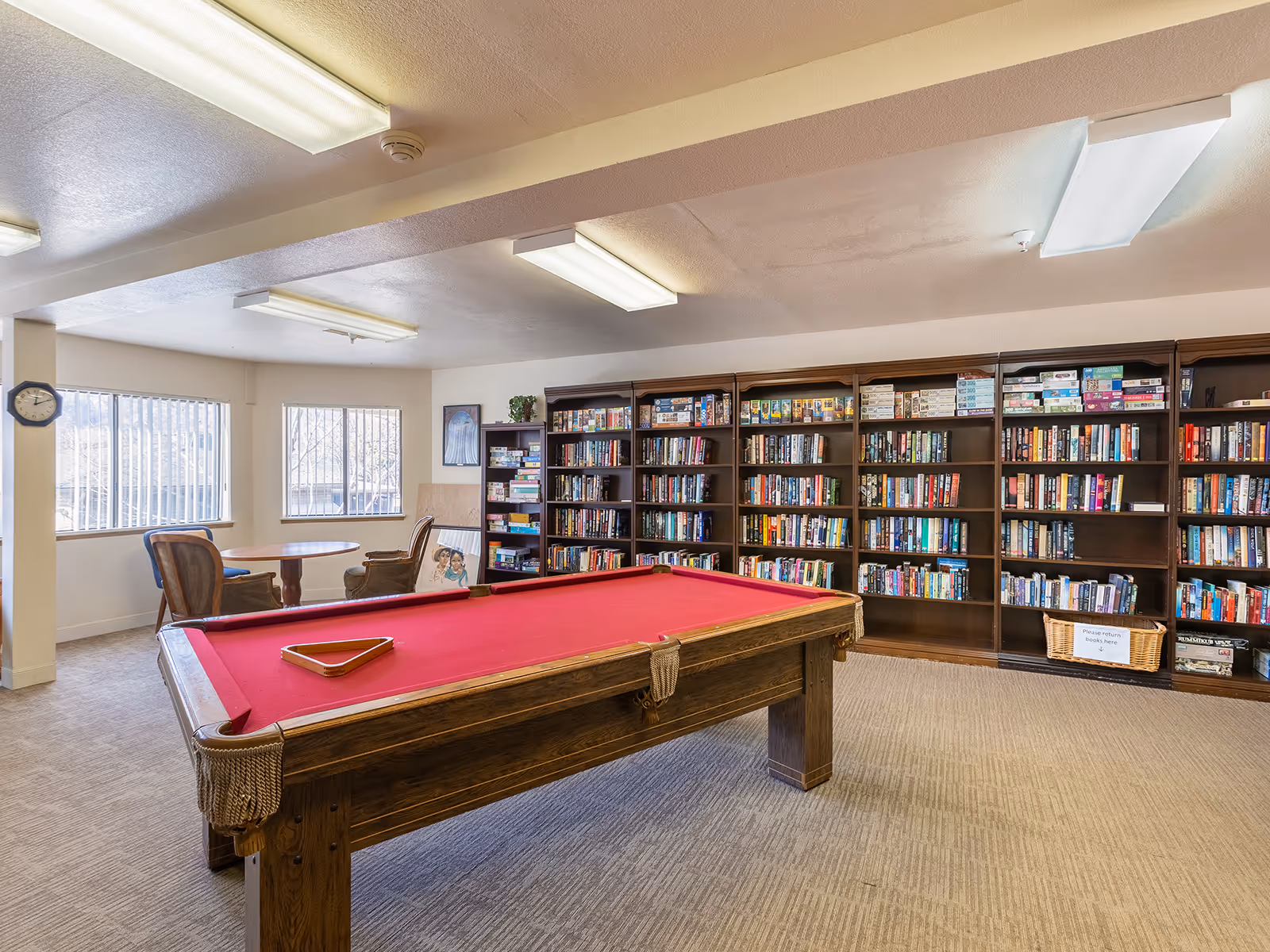 A recreational room with a red felt pool table in the center, surrounded by bookshelves filled with books and board games. There is a small round table with two chairs near two windows with vertical blinds, and a clock on the wall.