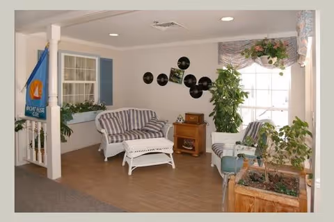 A cozy sitting area with white wicker furniture including a striped cushioned loveseat, an ottoman, and a chair. The room has light-colored walls decorated with vinyl records and a small wooden cabinet. There are several green plants and a large window with floral valance letting in natural light. A blue flag with a boat design hangs near a window with blue shutters.