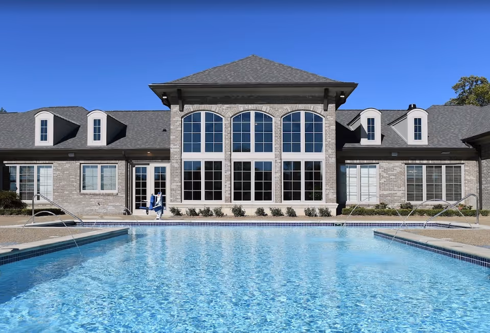 Outdoor swimming pool with clear blue water in front of a large brick building with multiple windows and a gray shingled roof under a clear blue sky.