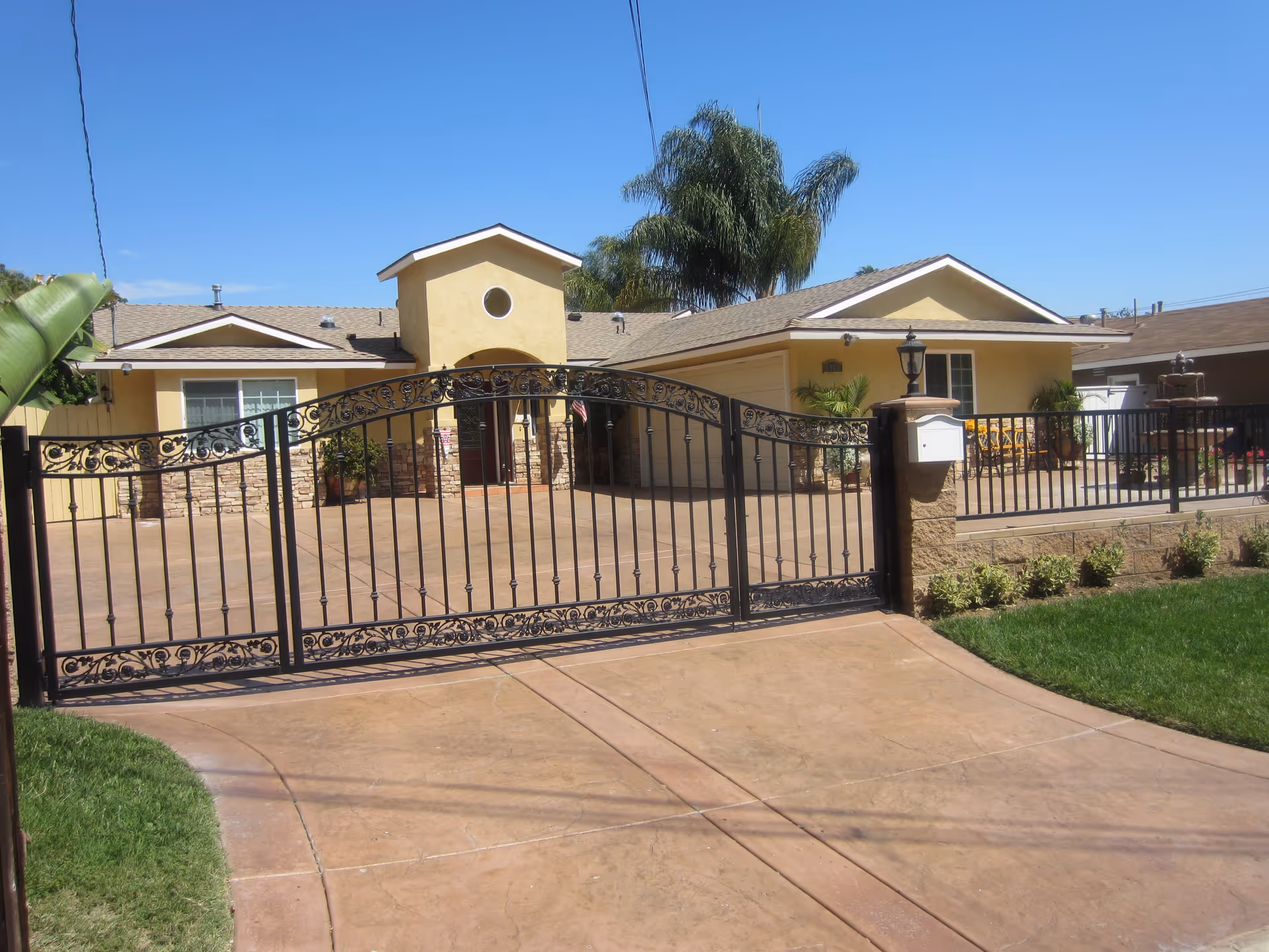 Front exterior view of a single-story yellow house with a decorative black metal gate, a driveway, and a small lawn with plants. The house has a garage, a front door with a small porch, and palm trees in the background under a clear blue sky.
