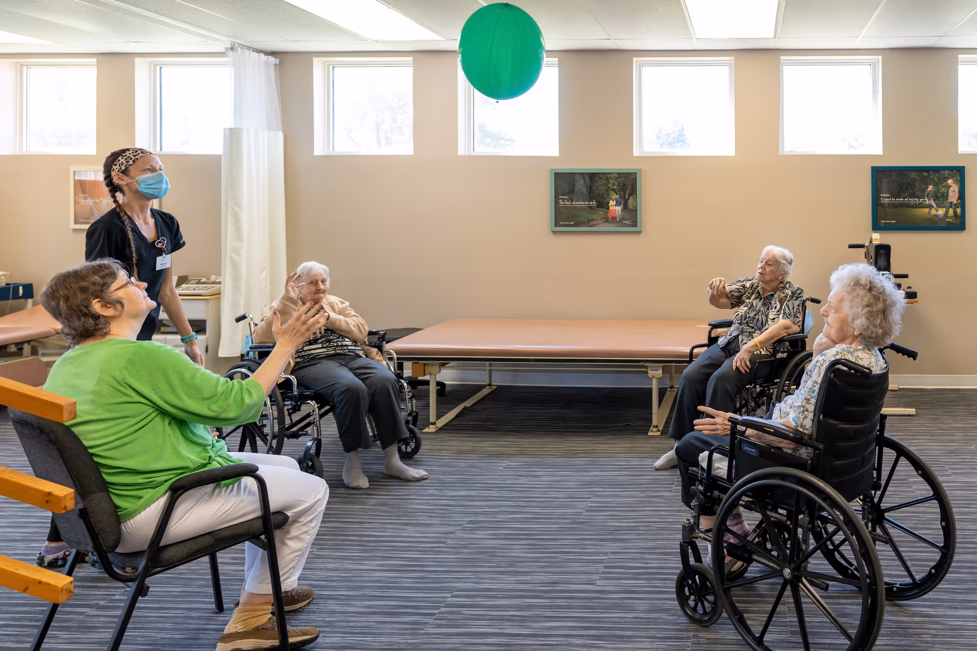 A group of elderly women, some in wheelchairs, and a caregiver wearing a mask are seated in a spacious room with large windows. They are playing with a green balloon, engaging in a light physical activity. The room has a padded therapy table against the wall and framed pictures hanging above it.