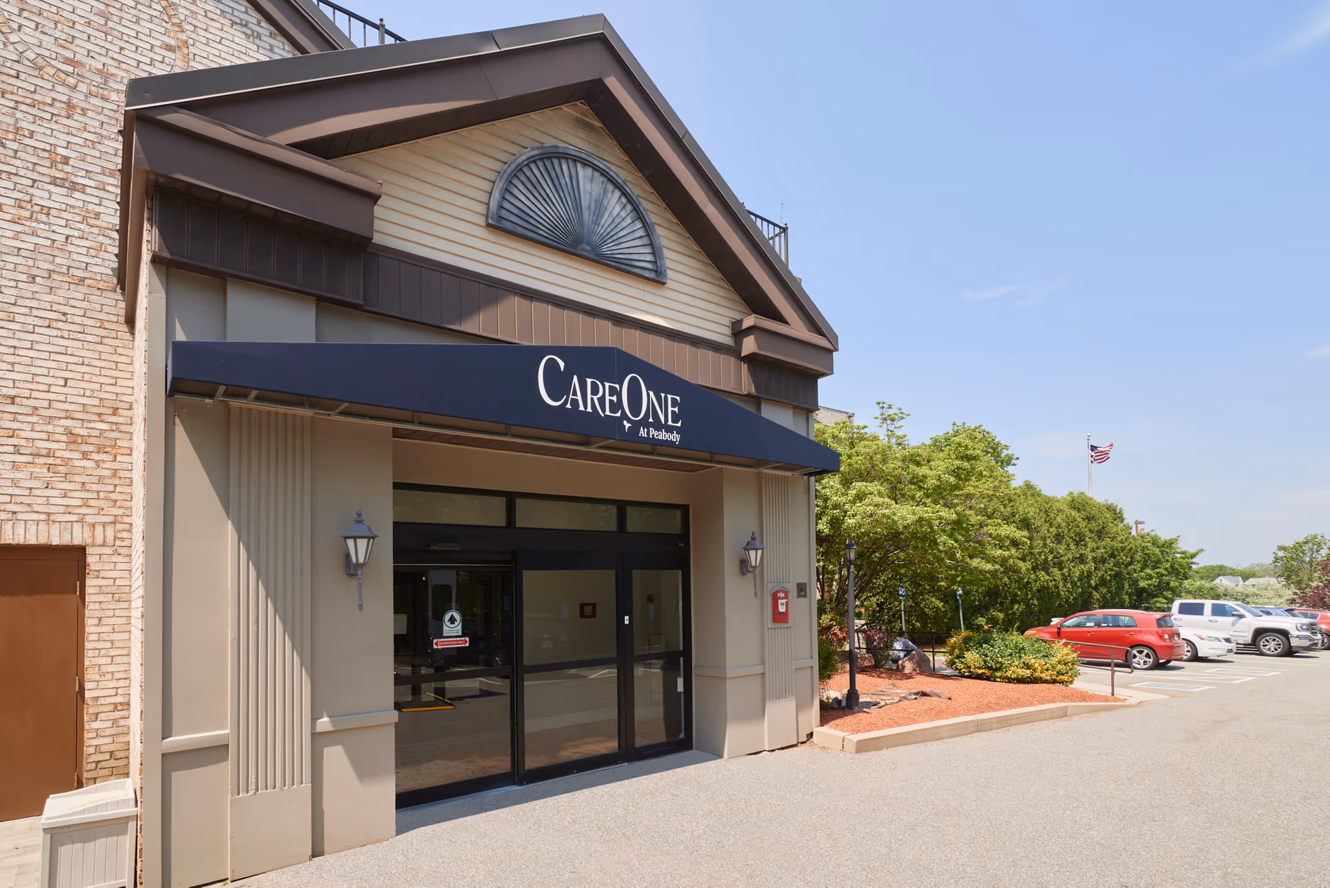 Entrance of CareOne at Peabody facility with a blue awning displaying the facility name above glass double doors. There are two wall-mounted lantern lights on either side of the entrance. To the right, there is a landscaped area with bushes and a parking lot with several cars under a clear blue sky.