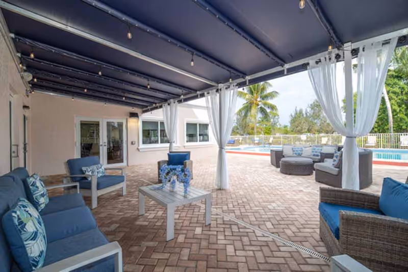 Covered outdoor patio area with blue cushioned wicker chairs and sofas arranged around white tables, sheer white curtains hanging from the ceiling, and a swimming pool visible in the background surrounded by palm trees and greenery.