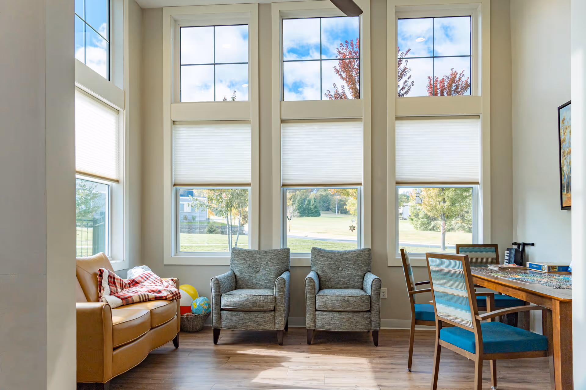 Bright seating area with tall windows, two gray armchairs, a tan sofa and a wooden table with teal chairs.