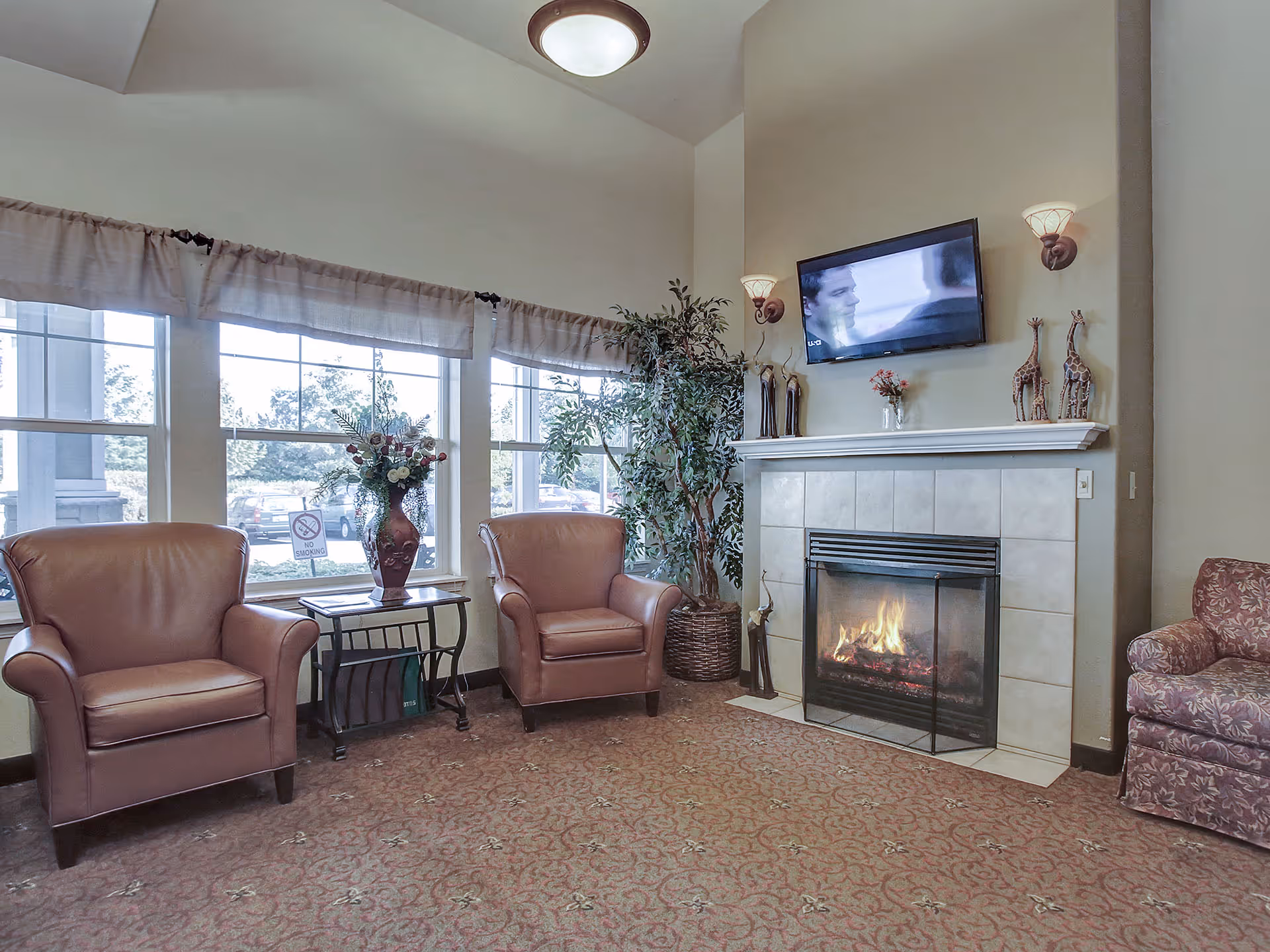 A cozy living room area with two brown leather armchairs and a floral-patterned armchair arranged around a fireplace with a fire burning. Above the fireplace is a mounted flat-screen TV. There are decorative items on the mantel, including giraffe figurines and a small vase with flowers. Large windows with light curtains allow natural light to fill the room, and a potted plant is placed next to the fireplace.