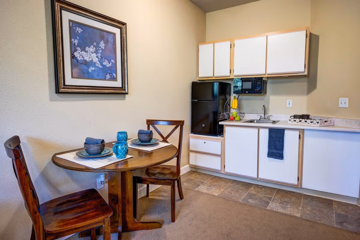 Small kitchenette area with white cabinets, a black mini refrigerator, a microwave, and a sink. Next to the kitchenette is a small wooden dining table set with two place settings, including blue bowls, plates, and glasses. A framed floral artwork hangs on the wall above the table.