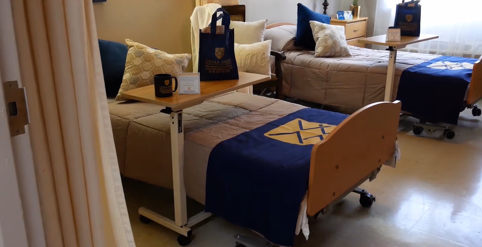 A room with two hospital-style beds, each with beige bedding and a blue blanket featuring a yellow emblem. Each bed has a wooden overbed table with a Cedar Ridge Health & Rehab Center tote bag, a mug, and a small sign. There are decorative pillows on the beds and a wooden nightstand between them with a plant and other items. The room has a light-colored floor and walls with a window letting in natural light.