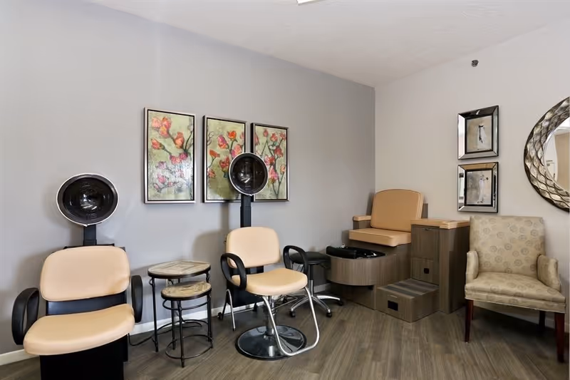 Salon-style room with beige hair-dryer chairs, a pedicure station, side tables, and floral wall art.