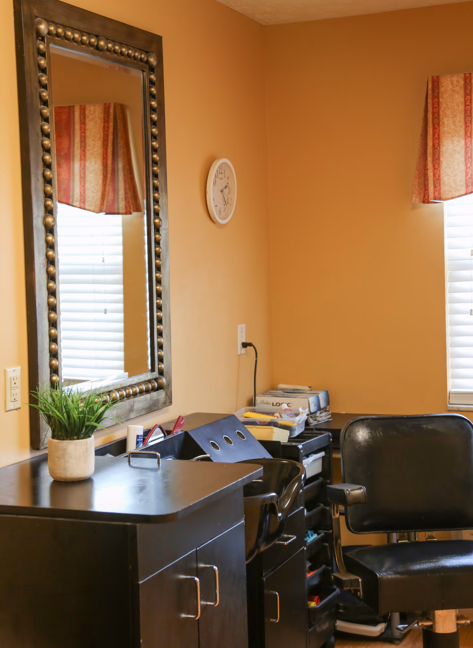 Interior room with tan walls featuring a large decorative mirror, a wall clock, two windows with red patterned valances, a black cabinet with a potted plant on top, and a black chair. There are various office supplies and papers on the cabinet and a rolling cart beside it.