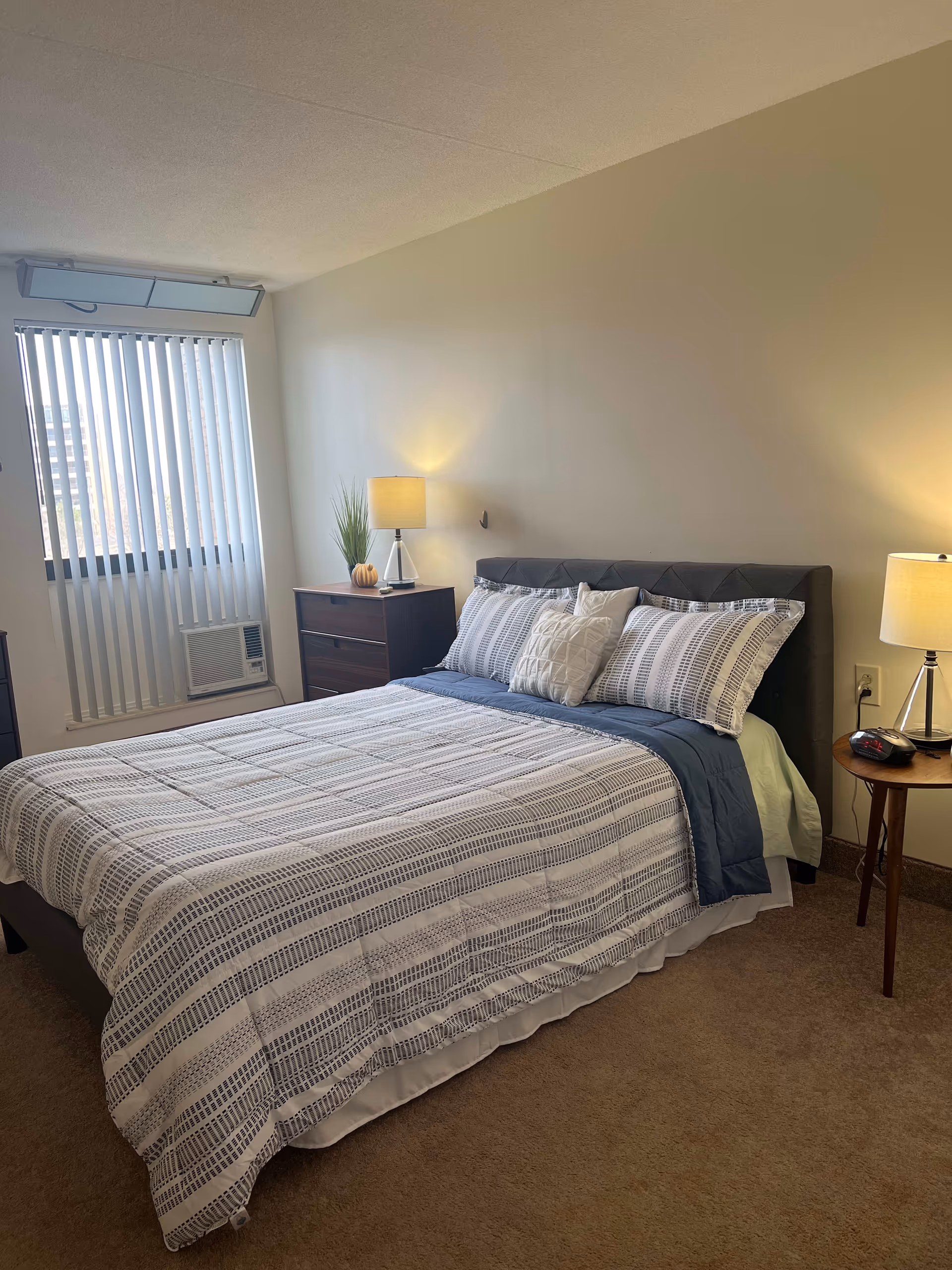 A neatly made bed with patterned bedding and multiple pillows in a bedroom. There are two bedside tables with lamps on either side of the bed. A window with vertical blinds and an air conditioning unit is visible on the left side of the room. The walls are plain and light-colored, and the floor is carpeted.