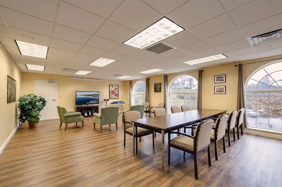 A spacious senior living common room with a long rectangular table surrounded by chairs on the right side and two green armchairs facing a flat-screen TV on the left side. The room has large arched windows with brown curtains, light yellow walls, wood flooring, and ceiling lights.