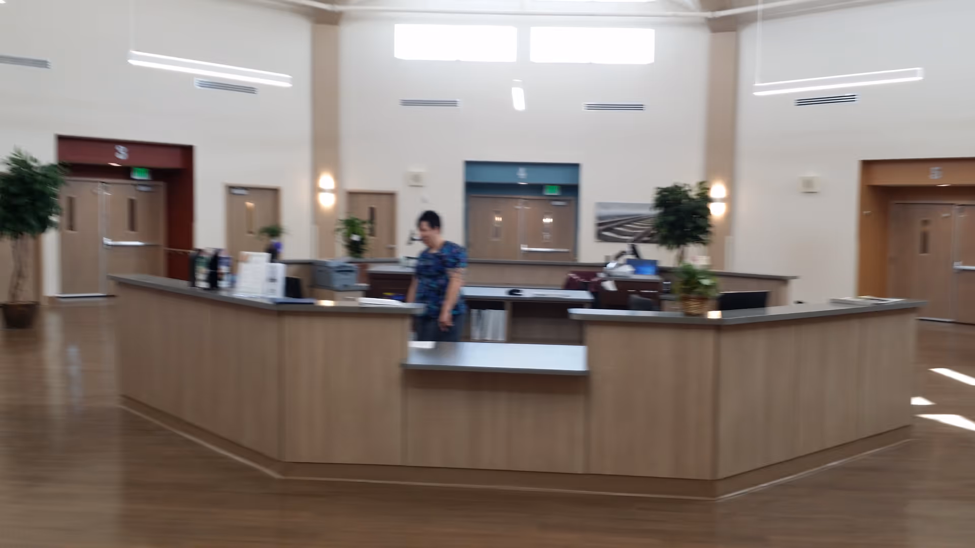A spacious reception area with a large wooden front desk in the center. A person is standing behind the desk. The room has high ceilings with bright lighting and several doors labeled with numbers. There are potted plants placed around the desk and along the walls.