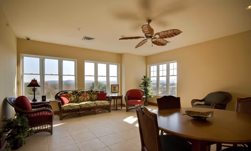 A bright living room with large windows letting in natural light. The room features a ceiling fan with leaf-shaped blades, a floral-patterned sofa with red cushions, two red armchairs, a wicker loveseat, a round wooden table with chairs, a floor lamp, and a potted plant. The walls are painted beige and the floor is tiled.