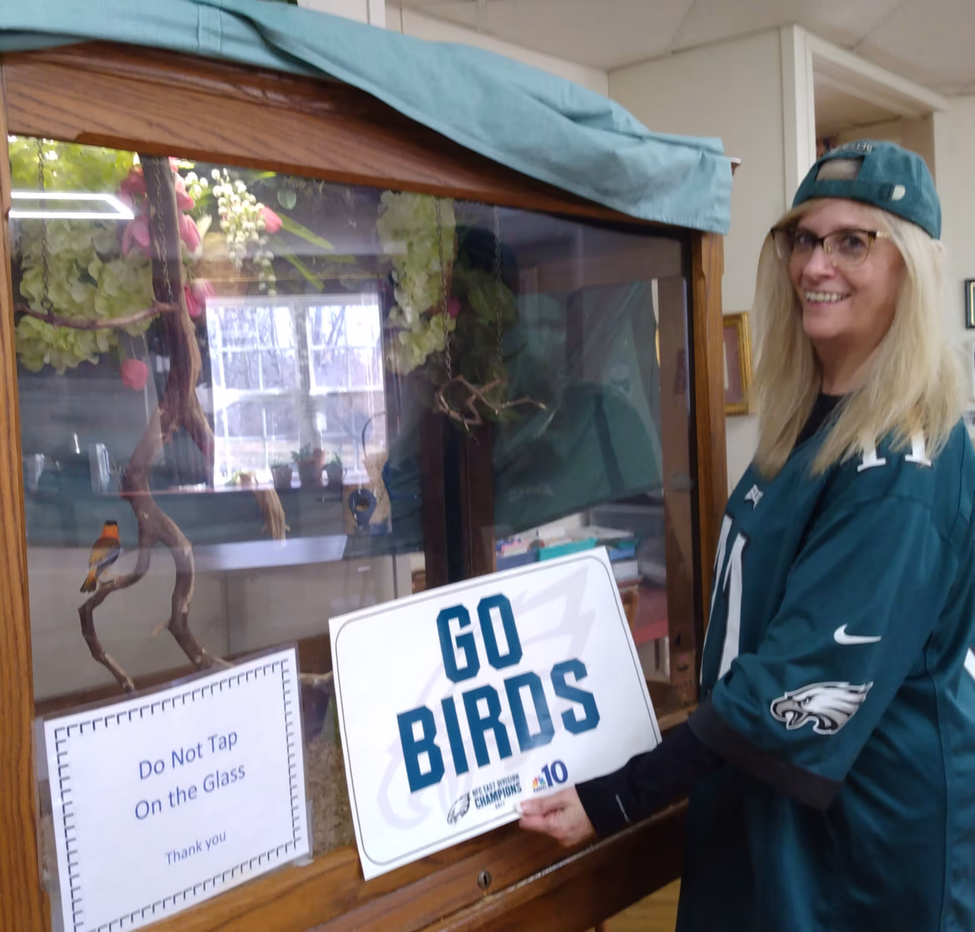 A woman wearing a Philadelphia Eagles jersey and a backwards cap is smiling and holding a sign that says 'GO BIRDS' in front of a glass enclosure with decorative branches and flowers inside. There is also a sign on the glass that reads 'Do Not Tap On the Glass Thank you.'