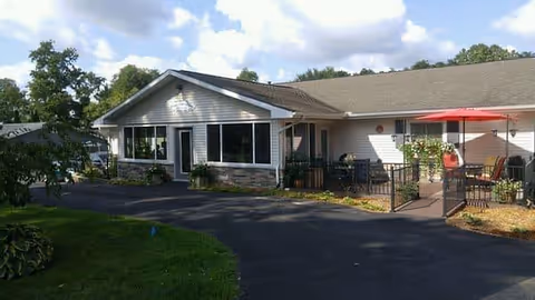 Single-story building with white siding and a gray roof, featuring large windows and a small fenced patio area with outdoor seating and a red umbrella. The building is surrounded by greenery and trees under a partly cloudy sky.