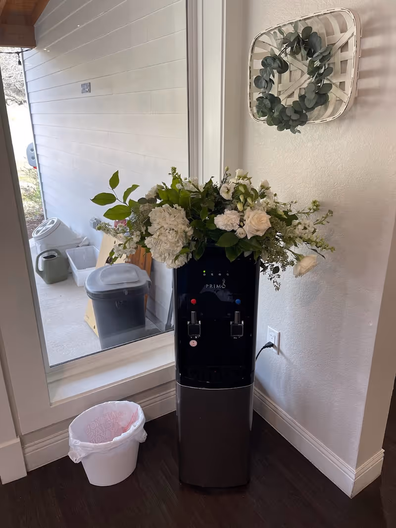 A black Primo water dispenser placed against a white wall with a floral arrangement on top. To the left, there is a large window showing an outdoor area with a watering can, trash bin, and other items. A small white trash can with a pink liner is on the floor next to the water dispenser. A decorative basket with green foliage hangs on the wall above the dispenser.