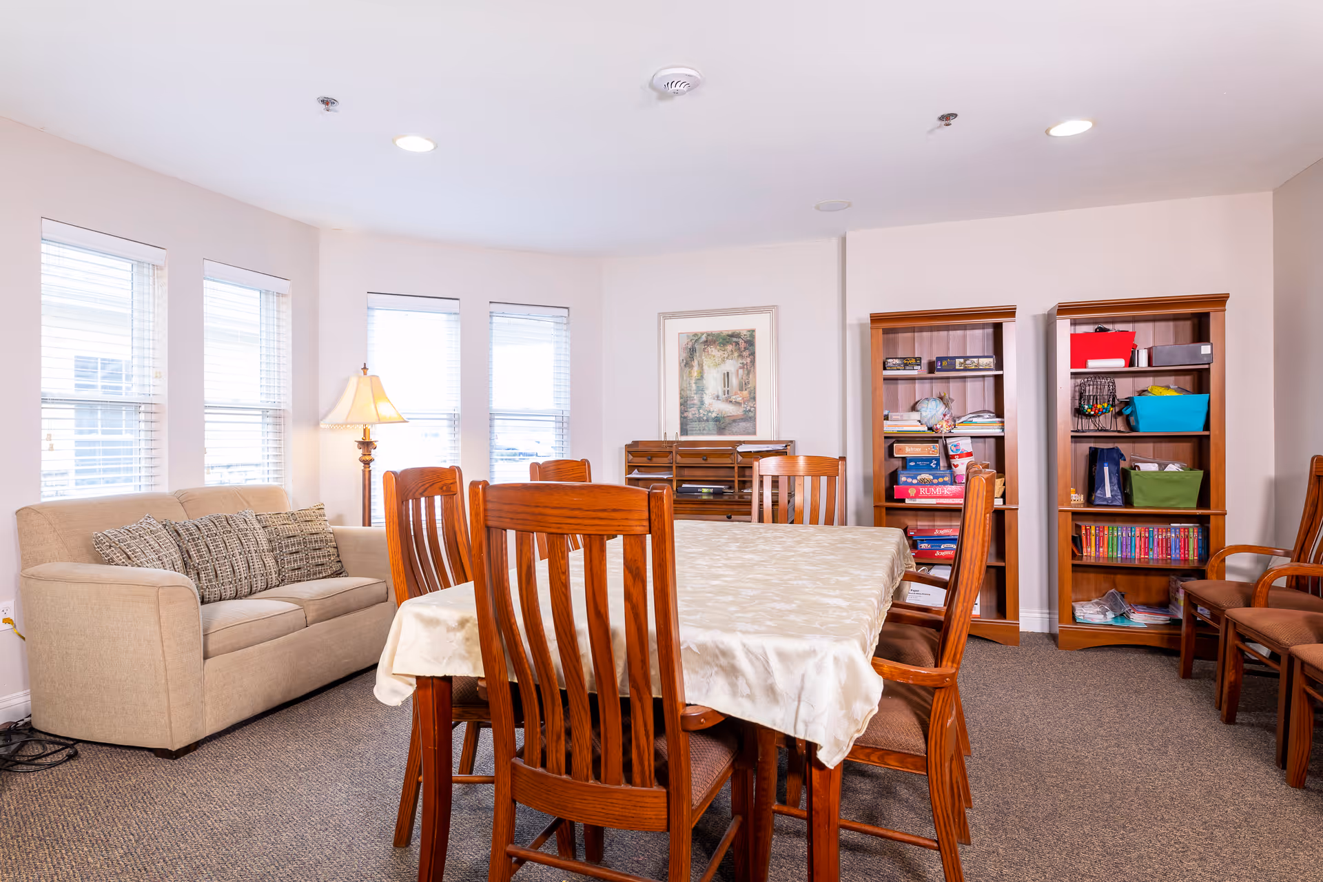A well-lit common room with a rectangular wooden table covered with a beige tablecloth surrounded by wooden chairs. To the left, there is a beige sofa with patterned cushions and a floor lamp beside it. The back wall features two wooden bookshelves filled with books, games, and storage bins. A framed painting hangs on the wall above a small wooden cabinet. The room has carpeted flooring and multiple windows letting in natural light.