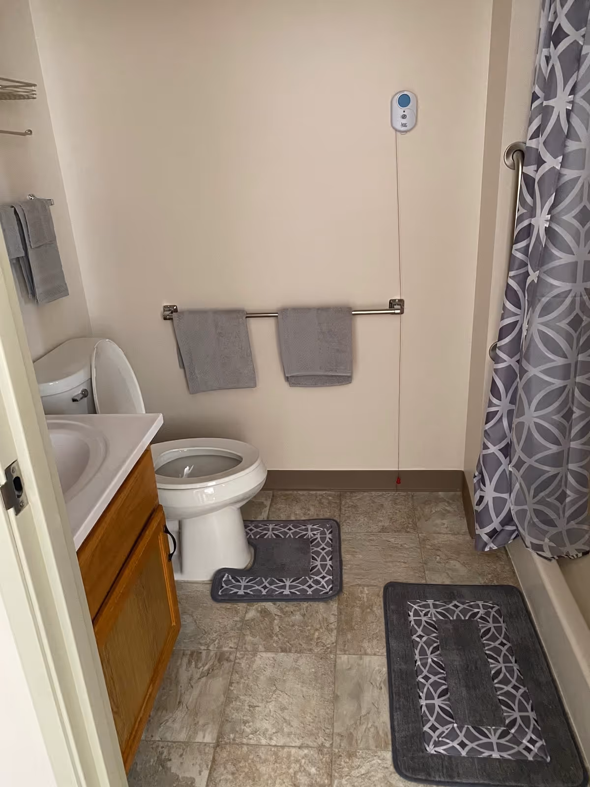 A small bathroom with a toilet, a wooden vanity with a white sink, a towel rack with two gray towels, a gray and white patterned shower curtain, and matching gray and white bath mats on a tiled floor. There is an emergency pull cord on the wall near the shower.