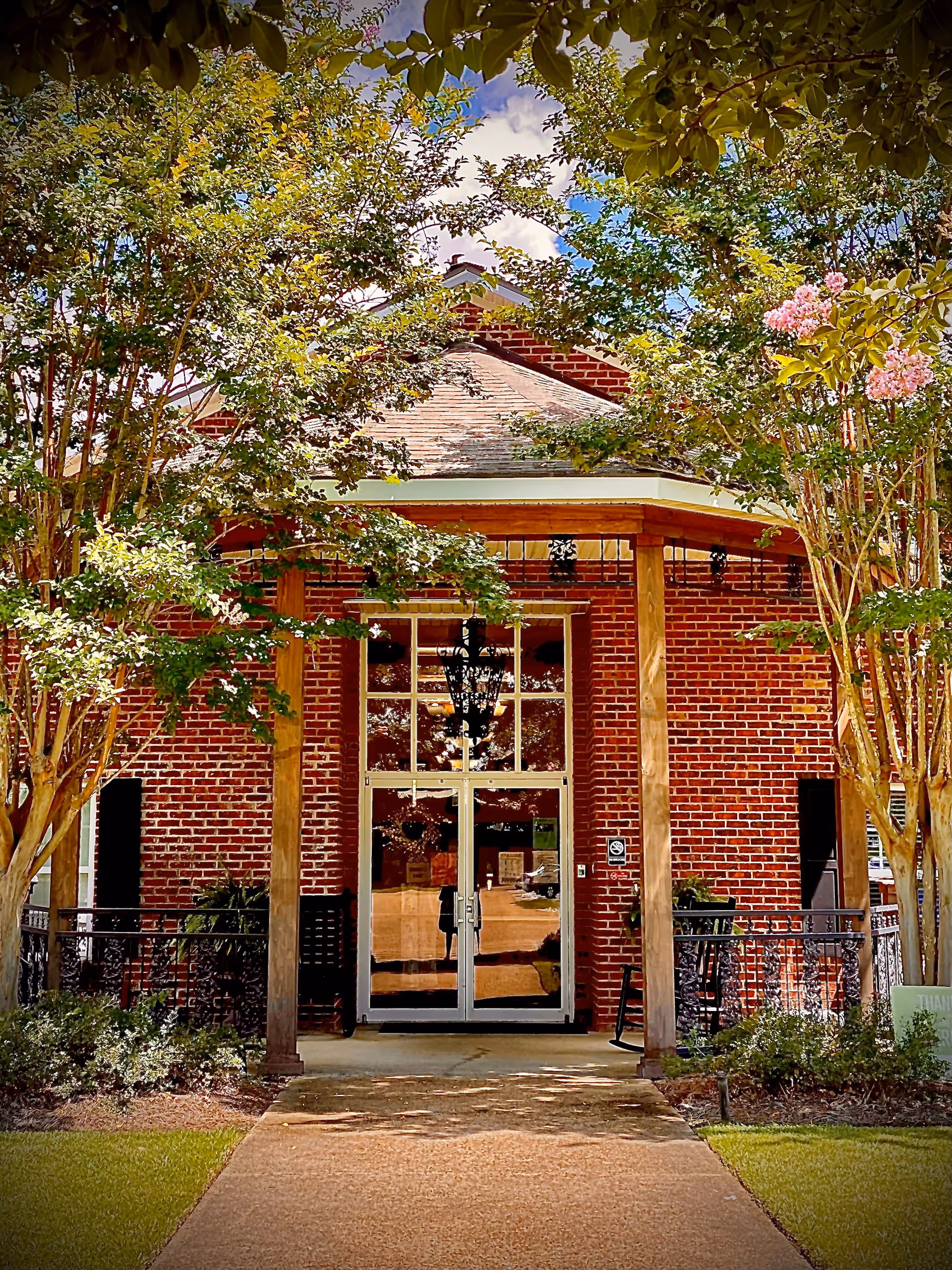 Front entrance of a brick building with large glass double doors, framed by wooden pillars and surrounded by green trees and shrubs under a partly cloudy sky.